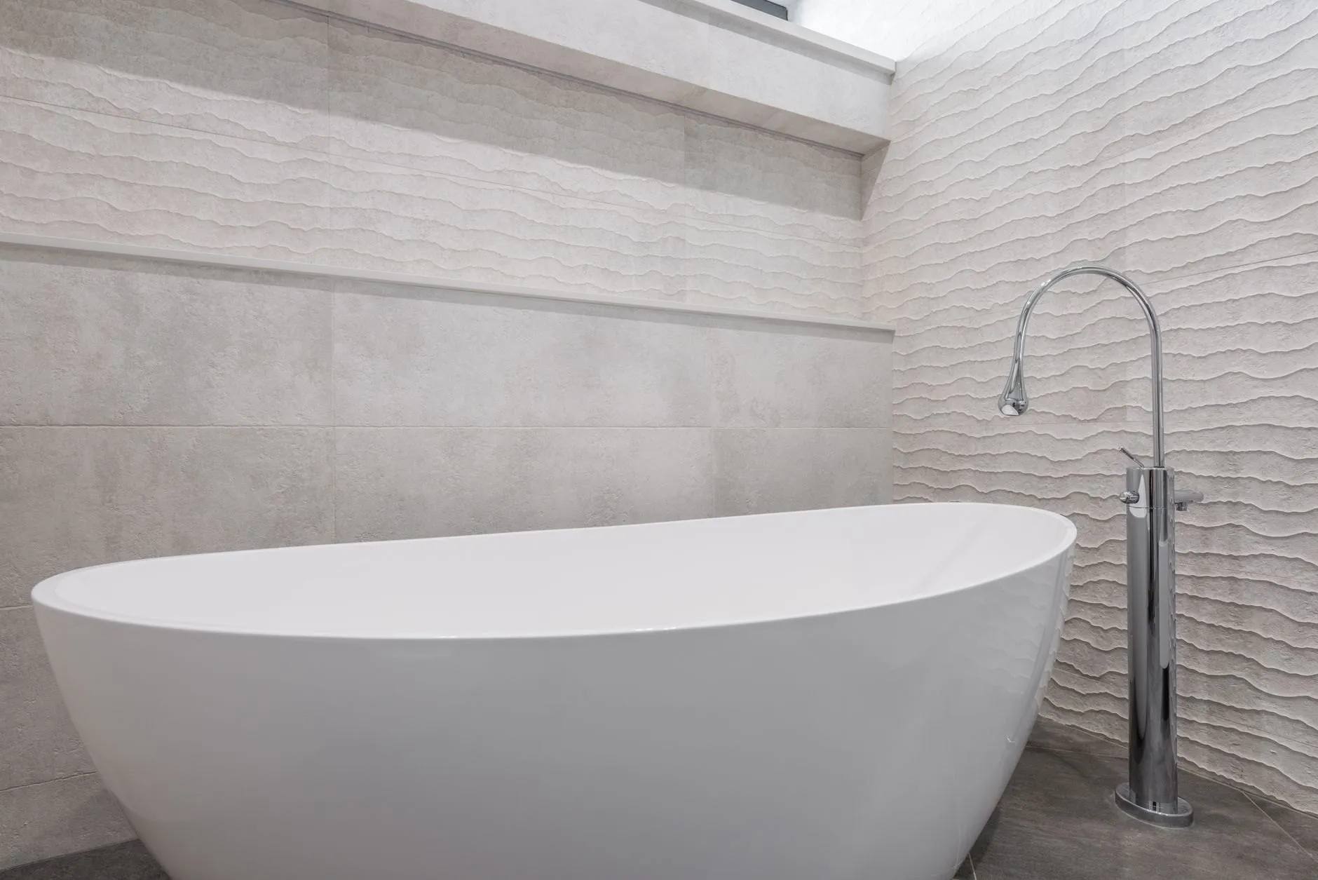 A freestanding white bath beside a floor-standing chrome bath filler against a wall of textured wave-pattern tiles