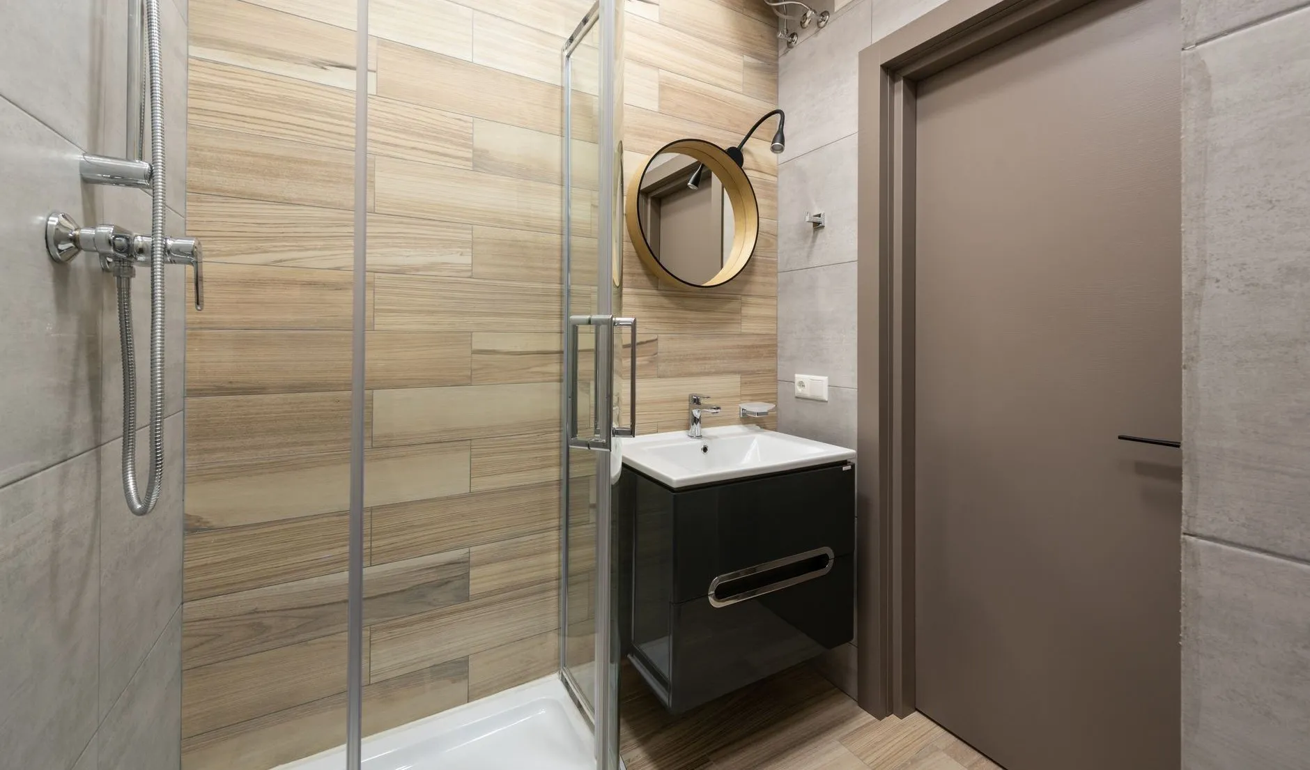 A small en-suite with a glass shower enclosure, oak-effect wall tiles, a round brass-framed mirror and a dark gloss vanity unit