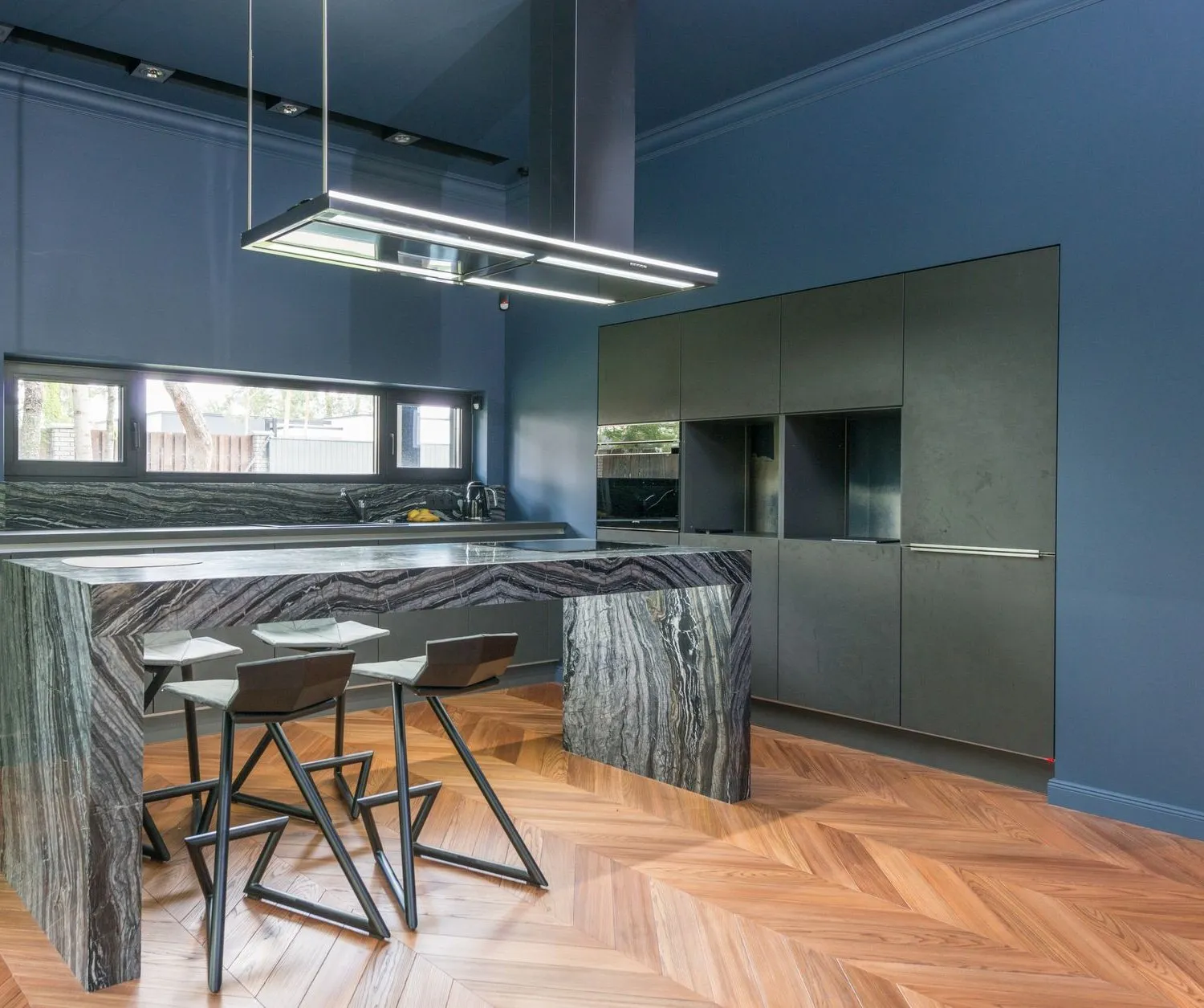 The finished kitchen: deep blue walls, a veined marble waterfall island with four stools, dark handleless units and a chevron oak floor
