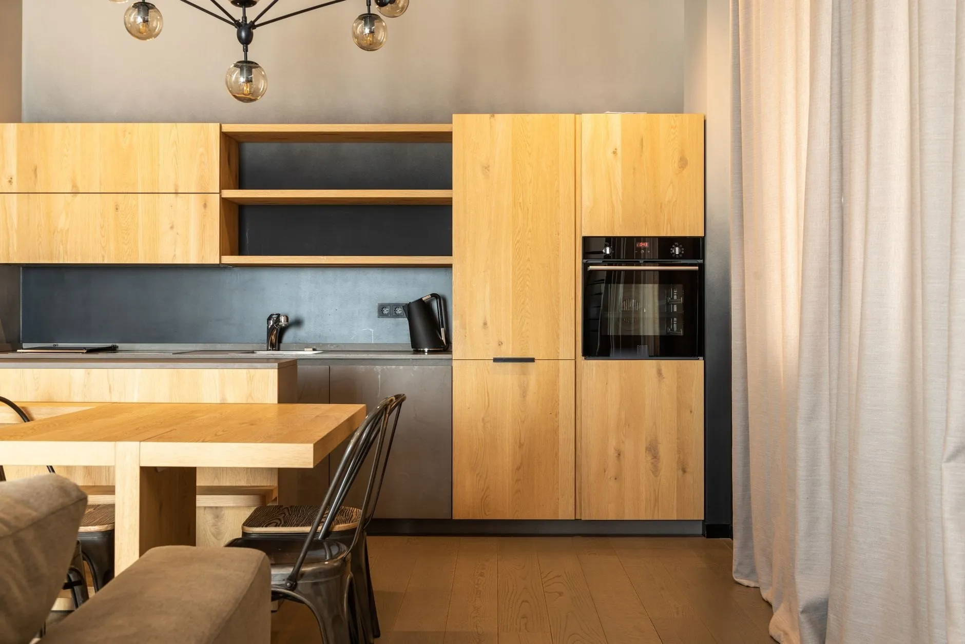 The finished kitchen: oak-effect units, open oak shelves over a charcoal splashback, a tall housing with a built-in oven and a wide oak table