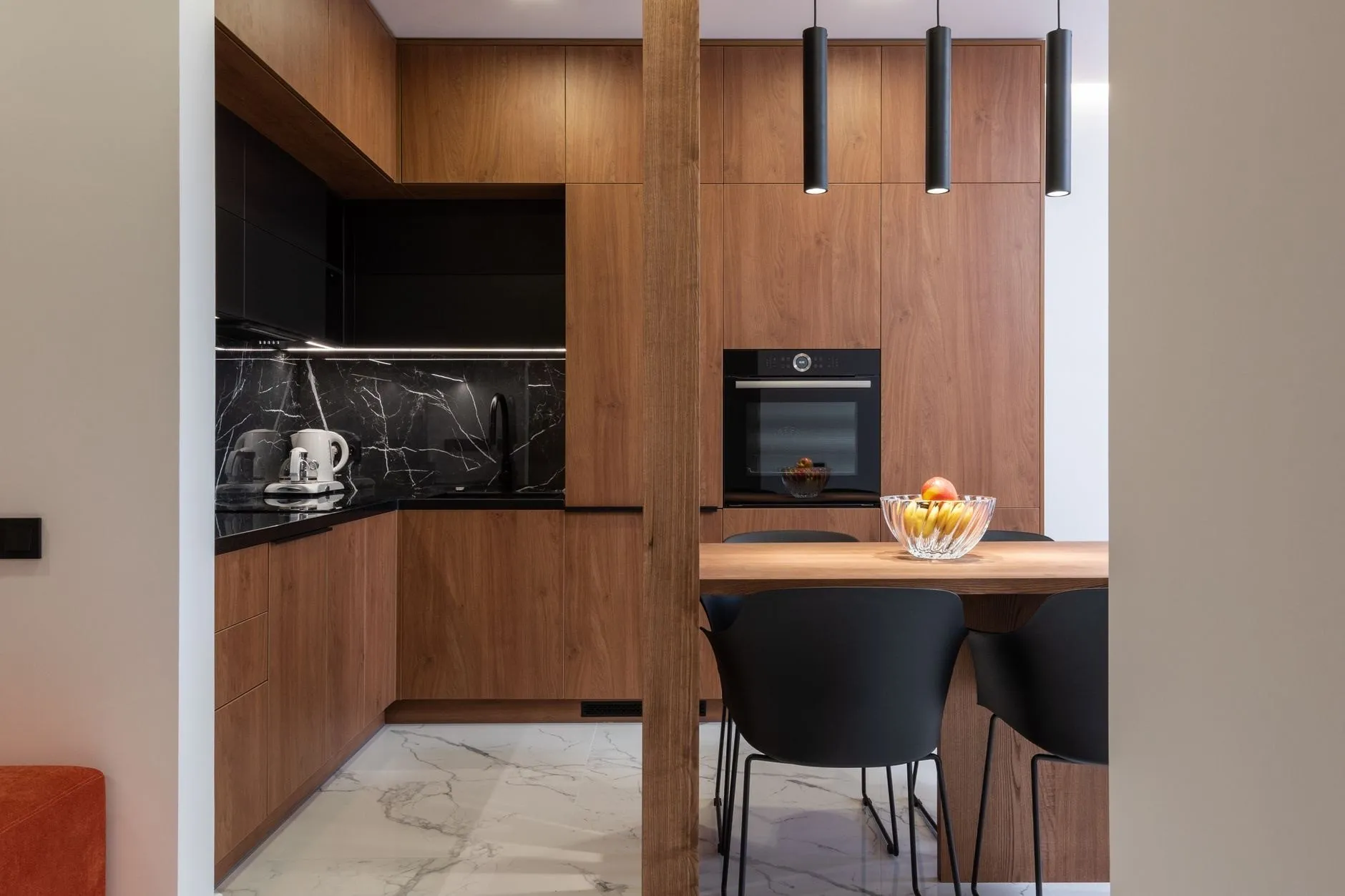 A walnut-veneer kitchen with a wall of tall units, a built-in oven, a black marble splashback and three slim black pendants over a table