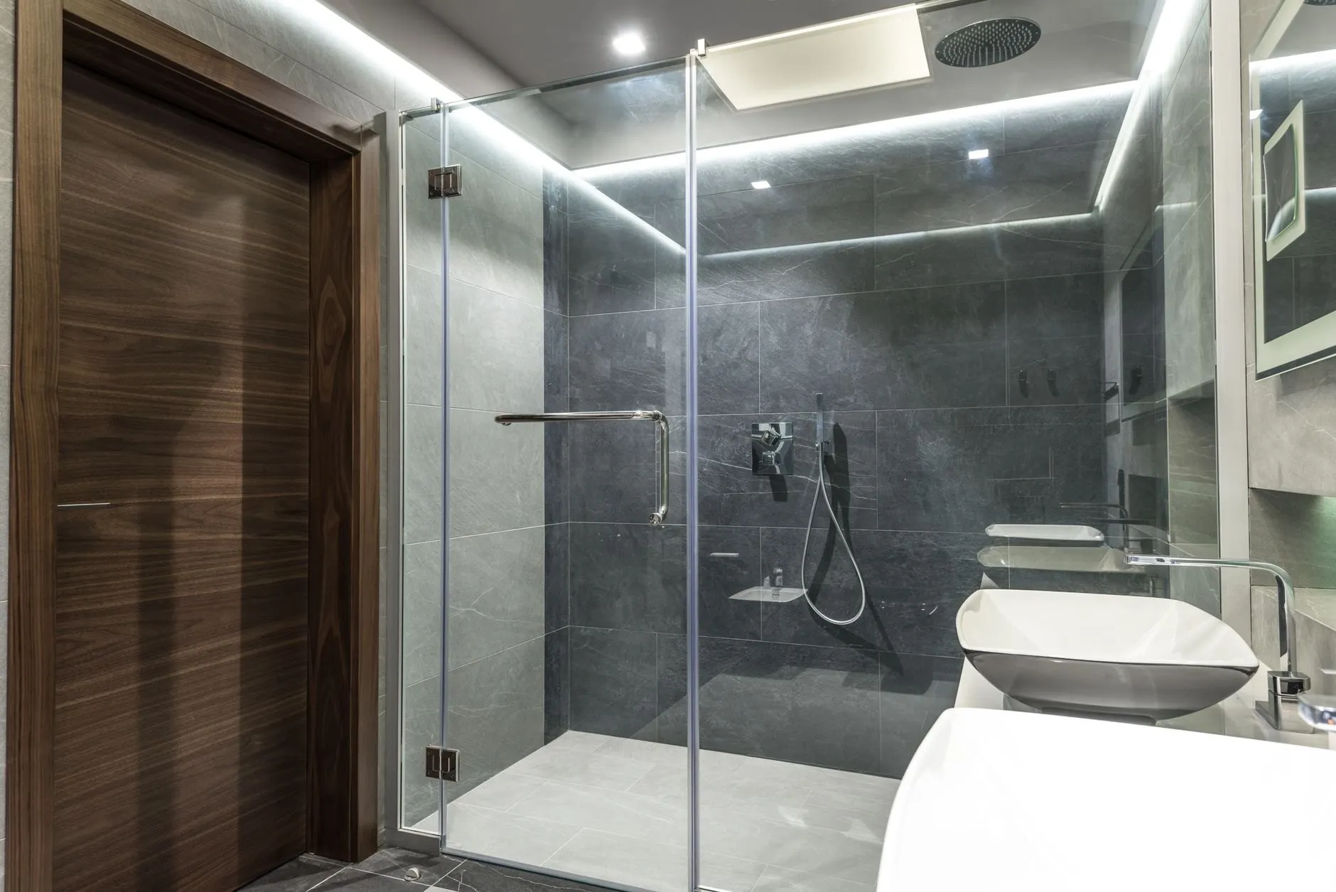 A frameless glass shower enclosure lined in charcoal stone tiles with a ceiling rain head and a walnut door beside it