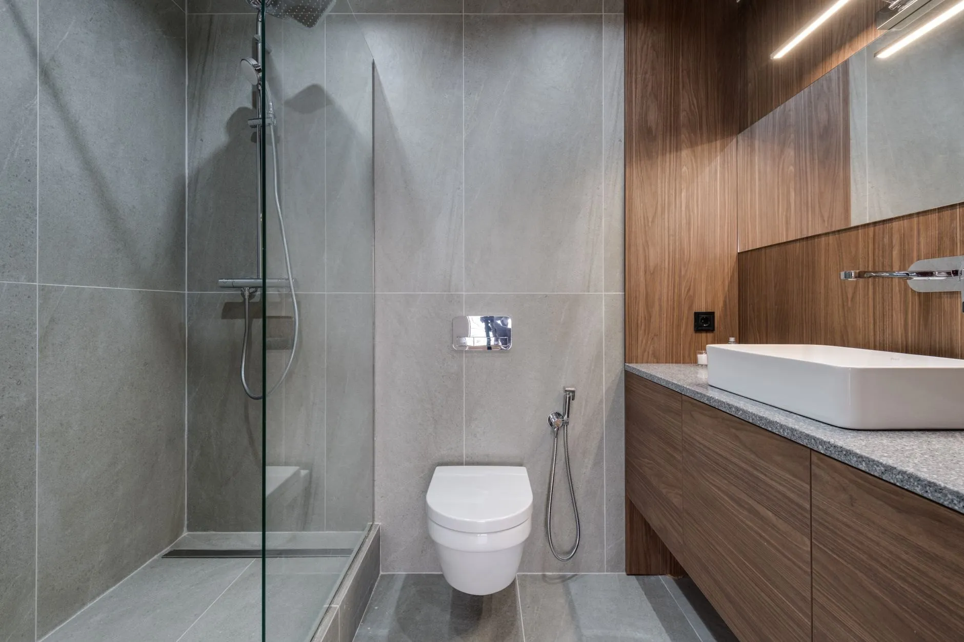 A walk-in shower with a single glass panel and grey stone-effect tiles beside a walnut vanity with a rectangular white basin