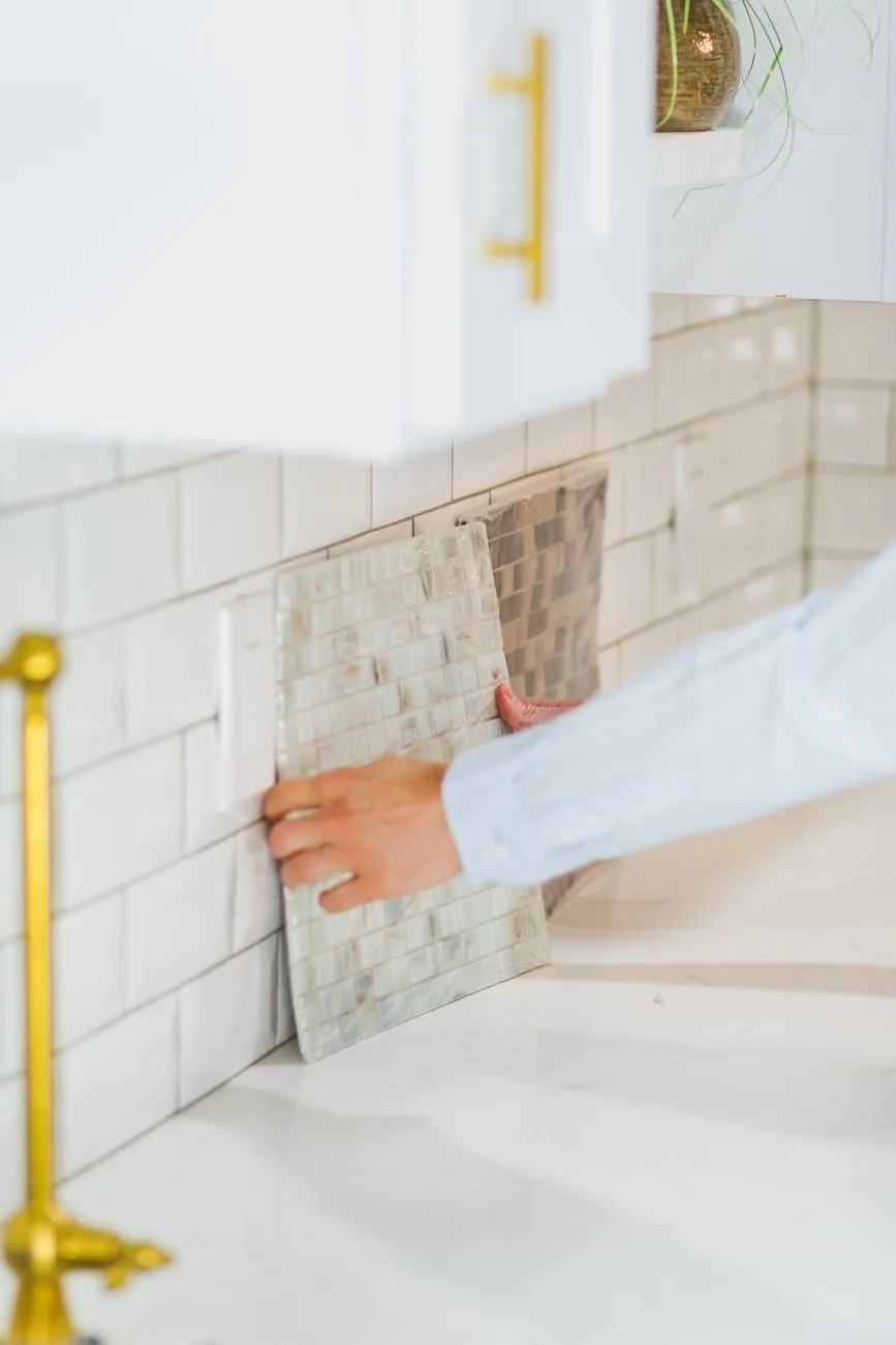 A hand pressing a mosaic tile sheet onto a white metro-tiled splashback under a painted wall unit with a brass handle